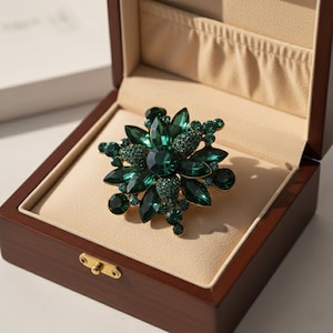 Puede incluir: Broche con forma floral con piedras preciosas de color verde esmeralda. El broche está engastado en metal dorado y se presenta en un joyero de madera con interior beige. El broche tiene una piedra central grande rodeada de piedras más pequeñas.