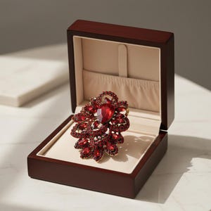 Può includere: Una spilla rossa ornata, con una pietra centrale a forma di cuore e gemme più piccole circostanti, presentata in un portagioie in legno scuro aperto con interno beige. La spilla ha una base in metallo dorato.