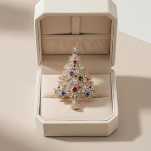 Peut inclure: Broche en forme de sapin de Noël en métal doré, incrustée de pierres précieuses claires et colorées. La broche est présentée dans un écrin à bijoux crème ouvert. Les pierres précieuses comprennent des accents rouges, bleus et verts.