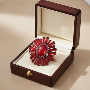Peut inclure: Une bague ornée d'une pierre rouge, de forme solaire, présentée dans un écrin en bois foncé. La bague est composée d'une pierre ovale centrale entourée de pierres rectangulaires. L'écrin a un fermoir doré.