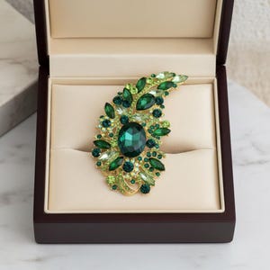 Peut inclure: Broche verte émeraude avec monture dorée, présentée dans un coffret en bois foncé. La broche présente une pierre centrale ovale entourée de cristaux et de strass verts de différentes tailles, formant un motif en forme de feuille.