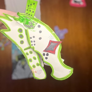 May include: A handmade paper craft in the shape of a green and white object with a domino-like piece on top. The object has a green outline and features black dots and a red diamond shape. The craft is held up by a hand.
