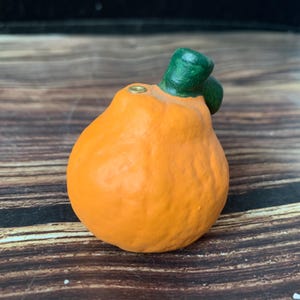 May include: A small, orange, fruit-shaped ceramic object with a textured surface and a dark green stem. A small, circular, metallic-colored opening is visible on the top of the orange body, positioned next to the stem, featuring a smooth finish.