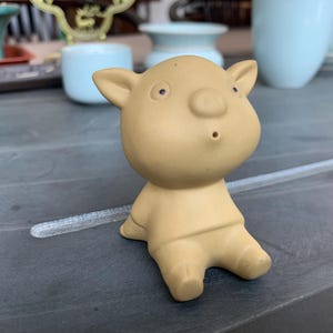 May include: A tan-colored ceramic figurine of a cartoon pig with small ears, a round nose, and a small mouth. The pig is sitting with its legs bent. The background includes light blue ceramic cups.