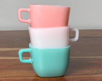 May include: A stack of three small glass mugs with square handles. The top mug is coral pink, the middle mug is opaque white, and the bottom mug is a bright turquoise blue. Each mug features a smooth, glossy finish and a simple, uniform shape.