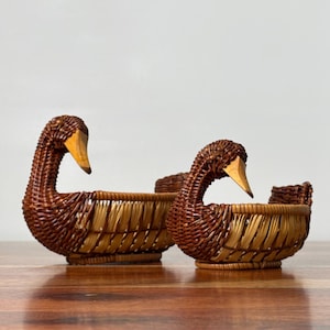 May include: Two woven baskets shaped like swans, crafted from dark brown and light tan wicker. Each basket features a smooth, light-coloured wooden beak and a curved neck, with a rounded body designed to hold small items within the woven structure.