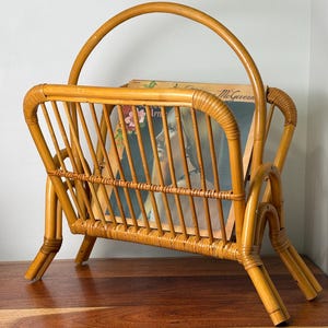 May include: A light brown rattan magazine rack with a curved handle and vertical slats. The rack holds a vinyl record sleeve featuring a portrait of a person with blonde hair and floral accents, with the text McGovern visible at the top.