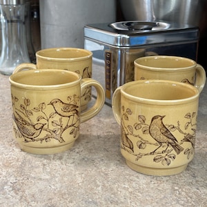 May include: Four beige ceramic mugs with brown bird and branch designs. Each mug has a handle and a slightly flared rim. The mugs are arranged on a countertop, suggesting a kitchen setting.