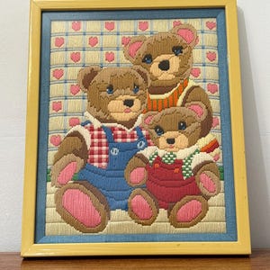 Peut inclure: Broderie encadrée représentant trois ours en peluche. Les ours sont bruns avec des yeux bleus et des détails roses. L'un porte une salopette bleue, un autre une écharpe rayée orange et blanche, et le troisième une salopette rouge. Le fond a un motif de cœurs.
