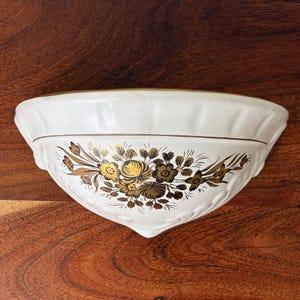Peut inclure: Une applique murale en céramique blanche en forme de demi-cercle ornée d'un motif floral doré sur le devant. L'objet présente un bord supérieur festonné avec un fin liseré doré, et le corps affiche un dessin botanique détaillé en or.