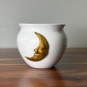 May include: A white ceramic planter featuring a raised, metallic gold crescent moon with a human face embossed on the front. The vessel has a rounded body with a flared rim and a smooth, glossy finish, showcasing a celestial design element.