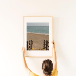 Könnte beinhalten: Gerahmter Druck einer Strandszene. Das Bild zeigt eine Holztreppe, die zu einem Sandstrand und dem Meer führt. Der Himmel ist bewölkt und das Wasser türkis. Der Rahmen hat eine helle Holzfarbe.