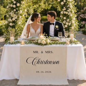 May include: A cream-colored fabric table runner featuring the black printed text MR. & MRS. Christenson and the date 08.16.2026. The runner is draped over a white tablecloth on a table set with plates, glassware, and white floral arrangements.