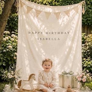 May include: A cream-colored fabric backdrop featuring a hanging bunting banner and the printed text HAPPY BIRTHDAY ISABELLA. The display includes a wooden rocking horse, a gift box with a sage green ribbon, and a wooden stacking toy on a rug.