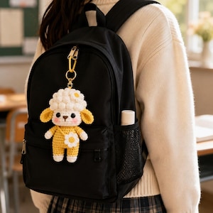 May include: A black backpack featuring a crocheted sheep keychain. The sheep has a white curly head, yellow ears, and a yellow body with a white flower detail. The keychain is attached to the top zip of the backpack with a gold-coloured ring.