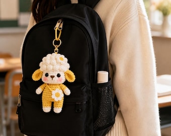 Puede incluir: Una mochila negra con un llavero de oveja tejido a ganchillo. La oveja tiene una cabeza blanca rizada, orejas amarillas y un cuerpo amarillo con una flor blanca. El llavero está sujeto a la cremallera superior con una anilla dorada.