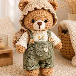 May include: A crocheted teddy bear wearing a tan sun hat, a cream-coloured long-sleeve shirt with small yellow flower embroidery, and olive green overalls with a white heart patch on the pocket. The bear also carries a small tan backpack.