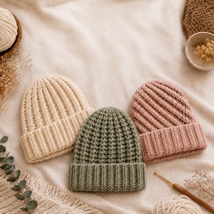 May include: Three knit beanies in cream, sage green, and dusty rose colors. Each hat features a thick, folded ribbed brim and a textured, vertical stitch pattern throughout the crown. The beanies are arranged side by side on a soft fabric surface.