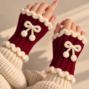 May include: A pair of fingerless gloves in a deep burgundy color with a ribbed knit texture. Each glove features a cream-colored crocheted bow appliqué and a scalloped, ruffled cream-colored trim along the top and bottom edges.