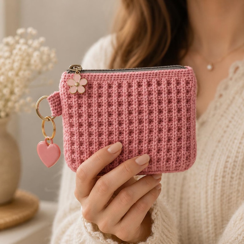 Puede incluir: Un monedero rectangular de ganchillo en color rosa empolvado con textura de punto de gofre. El tirador de la cremallera est&aacute; adornado con un peque&ntilde;o dije de flor de esmalte rosa claro y una anilla dorada con un colgante de coraz&oacute;n rosa.