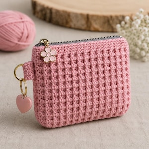 May include: A rectangular pink crochet coin purse featuring a textured waffle stitch pattern, a gray zipper closure, and a gold-tone metal ring attachment. The zipper pull is adorned with a small pink flower charm and a pink heart-shaped charm.