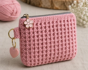 May include: A rectangular pink crochet coin purse featuring a textured waffle stitch pattern, a gray zipper closure, and a gold-tone metal ring attachment. The zipper pull is adorned with a small pink flower charm and a pink heart-shaped charm.