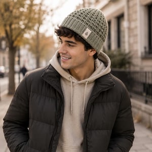 May include: A sage green knit beanie featuring a waffle-textured crown and a ribbed cuff. A small, rectangular off-white fabric tag is sewn onto the front of the cuff, displaying a simple dark brown graphic of a single leaf on a stem.