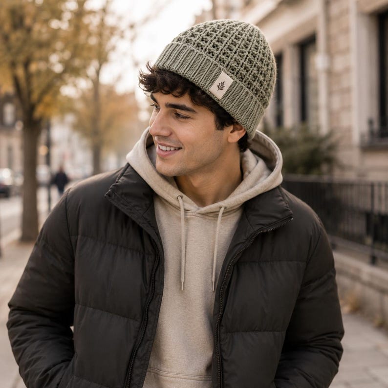 May include: A sage green knit beanie featuring a waffle-textured crown and a ribbed cuff. A small, rectangular off-white fabric tag is sewn onto the front of the cuff, displaying a simple dark brown graphic of a single leaf on a stem.