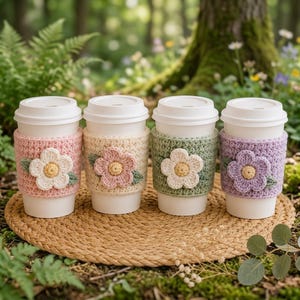 Op de afbeelding: Vier witte wegwerp koffiebekers, elk voorzien van een gehaakte hoes met textuur in pastelroze, crème, saliegroen en lavendel. Elke hoes is versierd met een enkele gehaakte bloem met een geel hartje en twee kleine groene blaadjes.