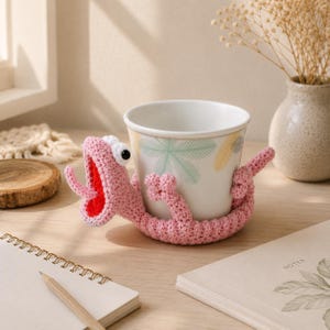 May include: A white ceramic mug featuring a light green and yellow palm leaf pattern, wrapped in a pink crocheted lizard cosy. The lizard has a wide-open mouth with a bright red interior, a long pink tongue, and two small white eyes with black pupils.