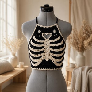 May include: A black crochet halter top featuring a cream-colored rib cage design on the front. The garment includes a heart-shaped cutout at the neckline and small star motifs above the rib cage, finished with a scalloped cream-colored border.