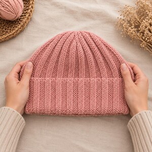 May include: A dusty rose pink beanie hat featuring a vertical ribbed knit pattern and a wide folded cuff. The hat is constructed from a thick, textured yarn and displays a consistent stitch throughout the crown and the brim of the garment.