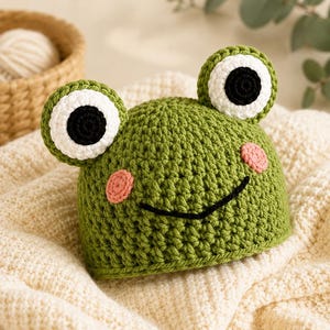 May include: A green crochet hat shaped like a frog, featuring two large eyes with white and black circular details, two small pink circular cheeks, and a black stitched mouth line across the front of the textured green yarn surface.