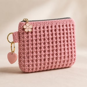 Puede incluir: Un monedero rectangular de ganchillo en color rosa con un patr&oacute;n de textura tipo gofre. El monedero tiene una cremallera gris oscuro, un dije de flor de metal dorado y un llavero dorado con un colgante rosa en forma de coraz&oacute;n.