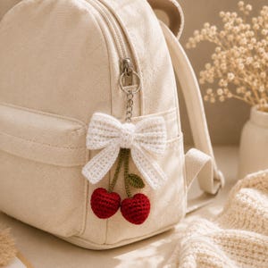May include: A cream-colored canvas backpack featuring a crocheted bag charm attached to the zipper. The charm consists of a white crocheted bow connected to two small, round, red crocheted cherries with green stems and a single green leaf.