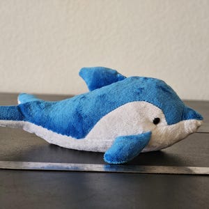 May include: A plush dolphin toy in shades of blue and white, with a black eye. The toy is approximately 12 inches long, with a ruler placed next to it for scale. The dolphin is lying on a dark surface.