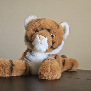 May include: A soft toy tiger with brown and black stripes, white details, and a tan nose. The plush animal is lying down on a dark surface, with its paws outstretched. The toy has a fluffy, furry texture.