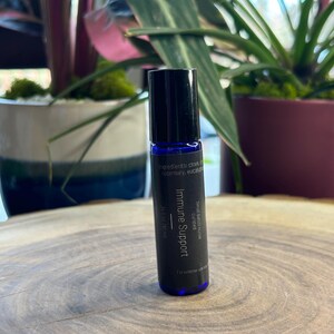 Op de afbeelding: Een kleine, donkerblauwe rollerfles met essentiële olie en een zwarte dop. Het etiket leest "Immune Support" en vermeldt ingrediënten. De fles is 10 ml.