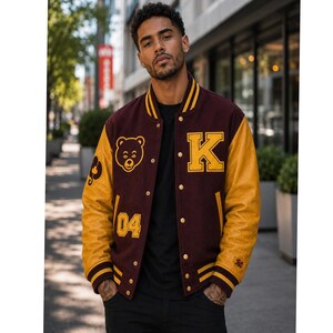 Può includere: Una giacca varsity con corpo in lana marrone e maniche in pelle gialla. Il davanti presenta una grafica con testa d'orso gialla, il numero 04 e una grande lettera K. La giacca ha una chiusura con bottoni a pressione e polsini a righe.