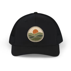 May include: A black trucker hat featuring a circular patch on the front. The patch displays a graphic of a bright orange sun setting over layered green hills, all enclosed within a cream-colored border against the solid black fabric of the cap.