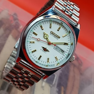 Könnte beinhalten: Eine silberfarbene Citizen Automatic Armbanduhr mit weißem Zifferblatt, goldenen Akzenten und einem roten Sekundenzeiger. Die Uhr zeigt den Tag und das Datum an. Das Armband besteht aus silbernen Gliedern.