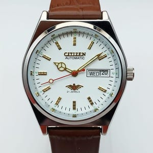Puede incluir: Un reloj de pulsera Citizen Automatic con esfera blanca, detalles dorados y correa de cuero marrón. El reloj muestra el día y la fecha y tiene la inscripción "21 Jewels". La caja del reloj es plateada.