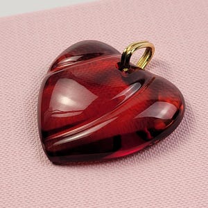 May include: A deep red, heart-shaped pendant with a smooth, translucent finish and subtle curved ridges. The pendant features a small, polished gold-tone metal bail attached to the top, designed for threading onto a chain or cord for jewelry.