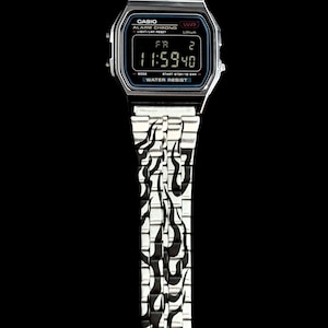 May include: A silver-tone digital wristwatch featuring a rectangular face with a black display showing the time 11:59 and the text CASIO ALARM CHRONO WATER RESIST. The metal link band is decorated with a black flame pattern throughout the entire length.