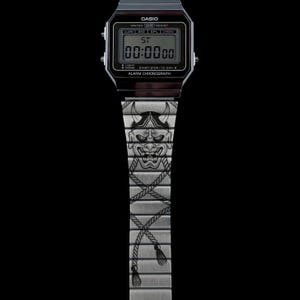 May include: A digital watch with a black face and a silver band. The band features a detailed engraving of a Japanese demon mask and rope design. The watch face displays "00:0000" and the brand name "CASIO".