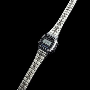May include: A silver Casio digital watch with a black screen displaying the time 9:49. The watch band features a repeating barbed wire design. The watch is set against a black background.