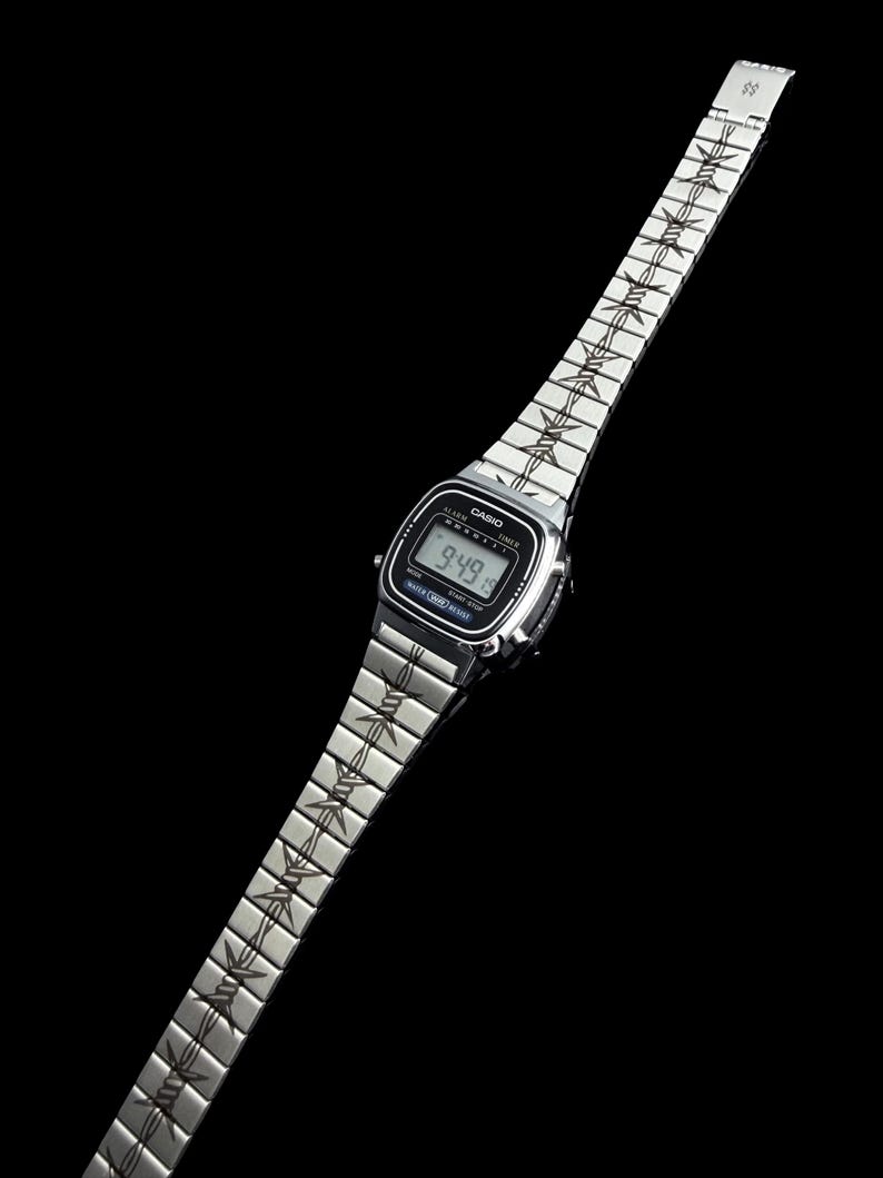 May include: A silver Casio digital watch with a black screen displaying the time 9:49. The watch band features a repeating barbed wire design. The watch is set against a black background.