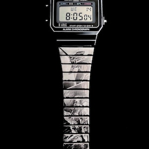 Könnte beinhalten: Eine schwarze Casio Digitaluhr mit silbernem Armband. Das Zifferblatt zeigt die Uhrzeit 8:05:04, den Wochentag und das Datum an. Das Armband hat ein Schwarz-Weiß-Design und den Schriftzug "EVANGELION".