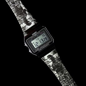 May include: Black digital Casio watch with a patterned band featuring a black and white design. The watch face displays the time and date. The band has a repeating pattern. The watch is set against a black background.