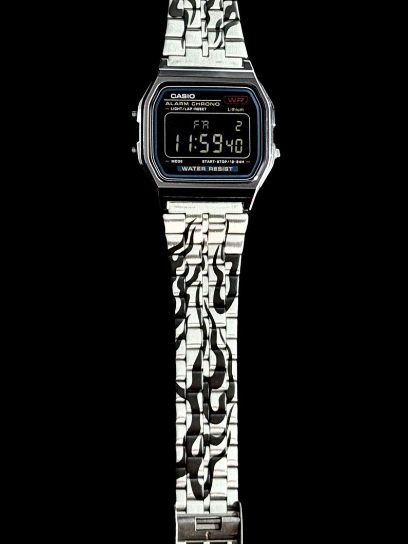 May include: A silver-tone digital wristwatch featuring a rectangular face with a black display showing the time 11:59 and the text CASIO ALARM CHRONO WATER RESIST. The metal link band is decorated with a black flame pattern throughout the entire length.